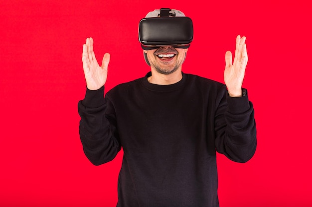 Mann mit Mütze in Schwarz mit Virtual-Reality-Brille, der lächelt, während er etwas virtuell berührt, auf rotem Hintergrund. Technologie-, VR-, Computer- und Hobbykonzept.