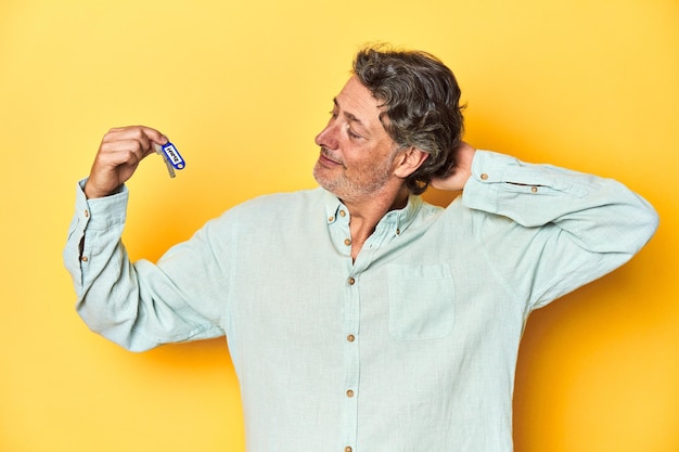 Foto mann mit neuen hausschlüsseln auf gelbem hintergrund berührt den rücken des kopfes und denkt und macht eine wahl