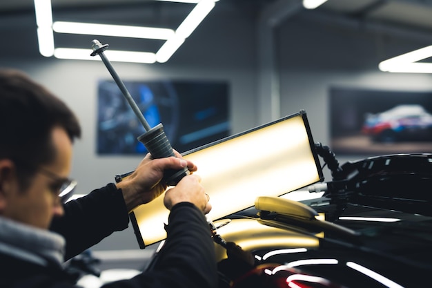 Mann mit professionellem Werkzeug und Lampe, um kleine Beule von der Rückseite eines schwarzen Autos in einer Garage zu entfernen Horizontale Innenaufnahme