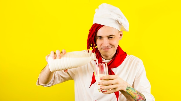 Mann mit roten Dreadlocks mit Flaschenmilch Selbstbewusster junger Koch in weißem Hemd und roter Krawatte gießt Milch aus einer Flasche in ein Glas vor gelbem Hintergrund