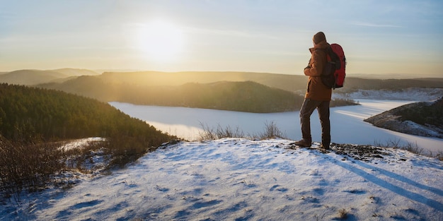 Foto mann mit rucksack steht auf dem hügel und genießt den sonnenuntergang. freiheit beim wandern im winter. panoramabanner.