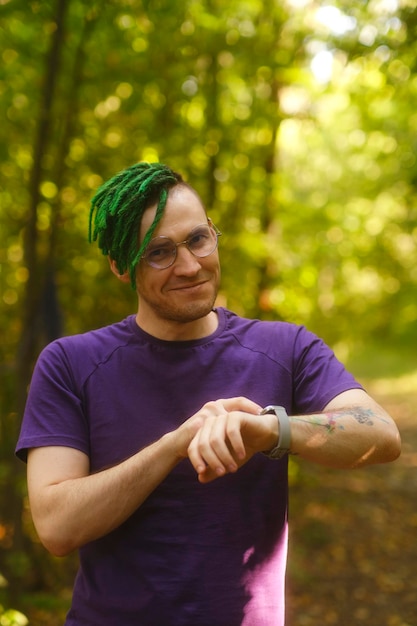 Mann mit Smart Watch im Park Mann in violettem T-Shirt mit Brille und grünen Deadlocks, der an Sommertagen im Park Benachrichtigungen auf Smart Watch überprüft