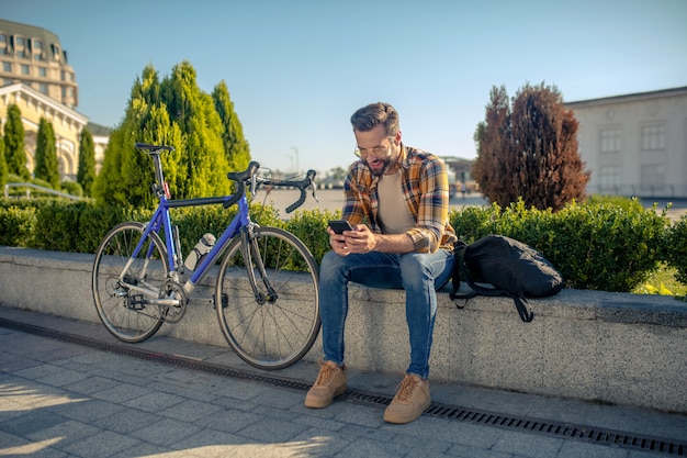 Mann mit Smartphone, das auf Straße nahe Fahrrad sitzt