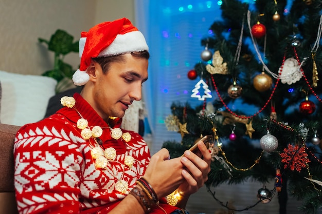 Mann mit Smartphone zu Weihnachten. Kerl feiert Neujahr allein zu Hause mit Weihnachtsmütze, Lichter am Weihnachtsbaum.