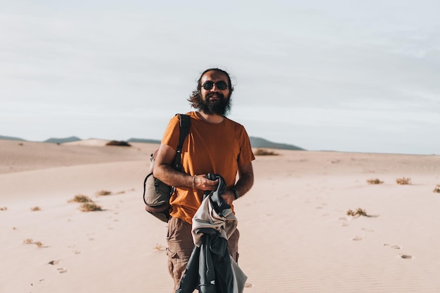 Mann mit Sonnenbrille schaut auf die Kamera, während er mit seinem Rucksack auf einer Schulter mitten in den Dünen lächelt