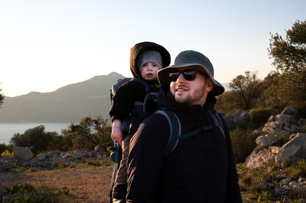 Mann mit Sonnenhut und Sonnenbrille mit BabyKleinkind im RucksackJunger gutaussehender Vater auf ReisenWandern mit Kind im Alter von 12 JahrenFamilienausflug in die NaturBergekaltes Wetter in der Nähe des Meeres