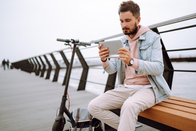 Mann mit Tablet, der Elektroroller am Pier in der Nähe des Meeres fährt Freiberufler Ökologischer Transport in der Stadt