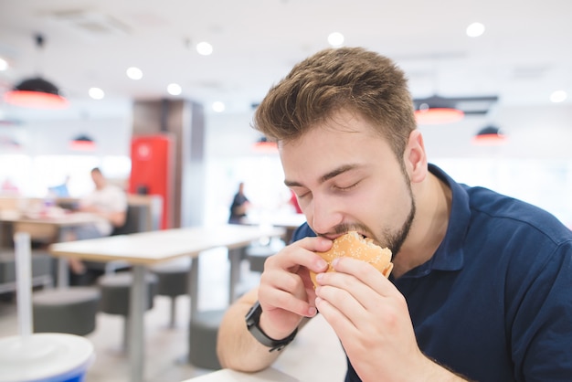 Mann mit Vergnügen beißt einen appetitlichen Burger auf einem Fast-Food-Restaurant