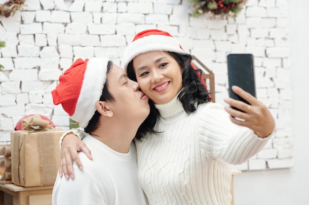 Mann mit Weihnachtsmütze, der Frau auf die Wange küsst, wenn sie ein Selfie auf dem Smartphone macht