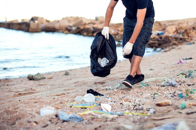 Foto mann mit weißen handschuhen und großem schwarzen paket, das müll am strand sammelt. umweltschutz- und planetenverschmutzungskonzept