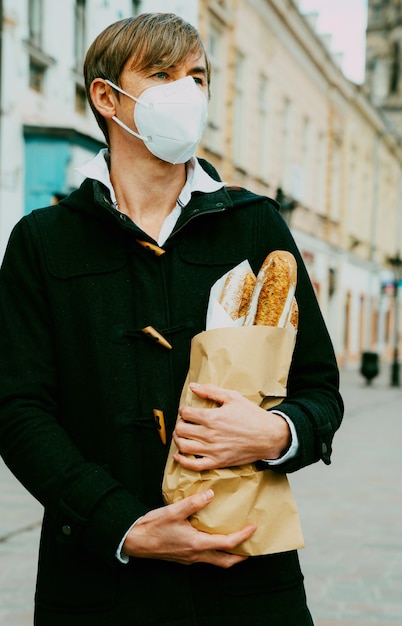 Mann mittleren Alters auf der Straße mit Brot, Baguette, Broteinkauf während der globalen Pandemie, Maske tragen, Brot aus der Bäckerei holen. Essen zum Mitnehmen, Bäckereieinkauf während Covid 19.