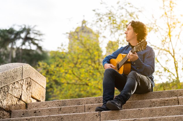 Mann mittleren Alters, der auf einer Treppe auf der Straße spanische Gitarre spielt. Er trägt Jeans und blaues Hemd