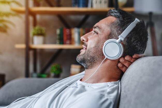 Mann mittleren Alters, der Musik mit Kopfhörern hört, entspannt auf dem Sofa zu Hause