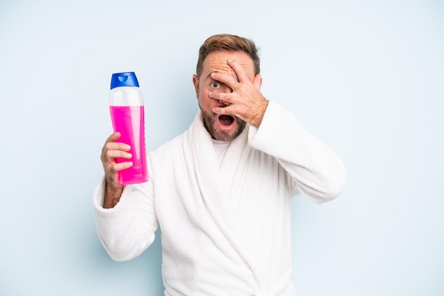 Mann mittleren Alters, der schockiert, verängstigt oder verängstigt aussieht und das Gesicht mit der Hand bedeckt. Shampoo-Flasche