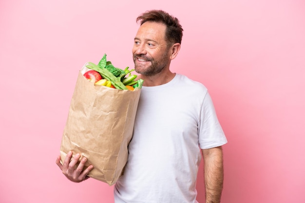 Mann mittleren Alters hält eine Einkaufstasche isoliert auf rosa Hintergrund, schaut zur Seite und lächelt