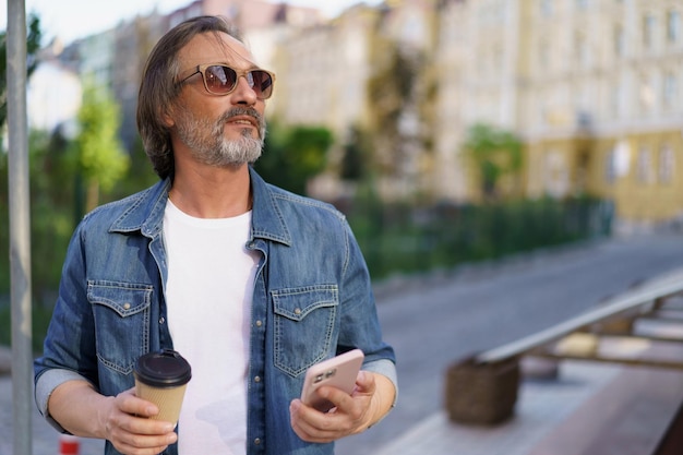 Mann mittleren Alters ließ sich vom Handy ablenken und genoss beim Kaffeetrinken im Freien den Sonnenuntergang. Mann mittleren Alters blickte weg auf die Straße, während er seine freie Zeit von der Arbeit genoss. Geschäftlich unterwegs