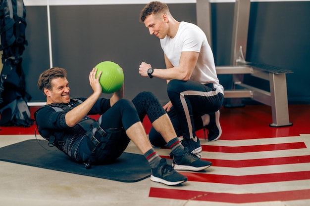 Mann mittleren Alters macht Sit-up-Übungen während des EMS-Trainings mit Personal Trainer im Fitnessstudio.