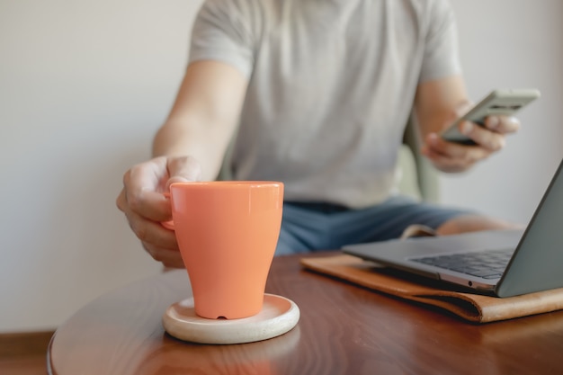 Mann nimmt eine orange Kaffeetasse auf, während er an seinem Laptop arbeitet.