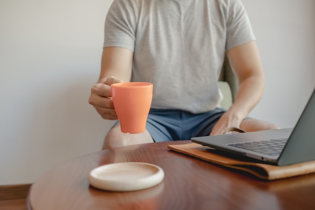 Mann nimmt Kaffeetasse auf, während er an seinem Laptop arbeitet.