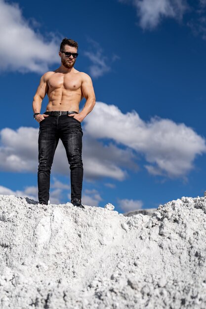 Mann ohne Hemd steht auf weißem Felsen Kerl in coolen Jeans hält die Hände in den Taschen Blauer Himmel mit flauschigen Wolken