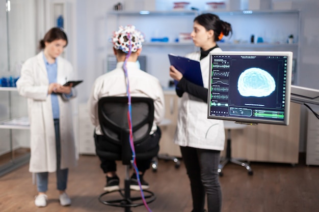 Mann-Patient, der ein leistungsfähiges EEG-Headset trägt, das die elektrische Aktivität des Gehirns im neurologischen Forschungslabor scannt, während ein medizinischer Forscher es kontrolliert, das Nervensystem untersucht, eine Krankenschwester macht Notizen