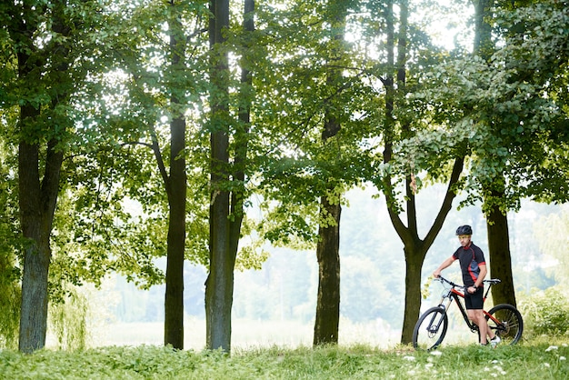 Mann Radfahrer im professionellen Kleidungsstück, das nahe Fahrrad in schönem sonnigem grünem Park unter hohen Bäumen steht