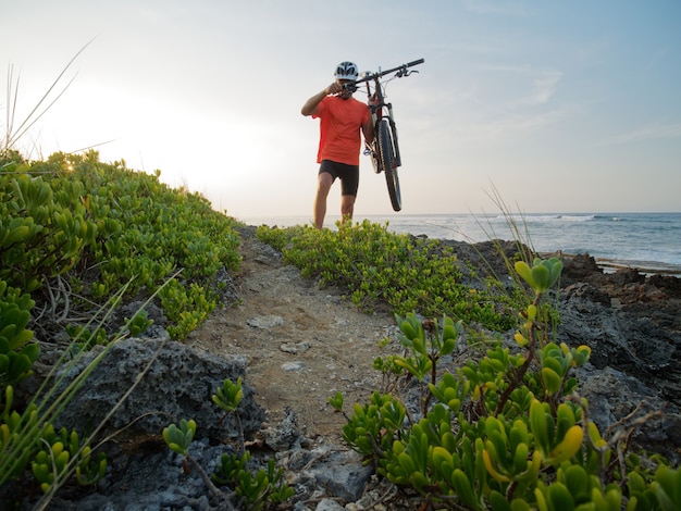 Foto mann-radfahrer trägt ein fahrrad in seinen händen, an der küste des ozeans. felsstrand.