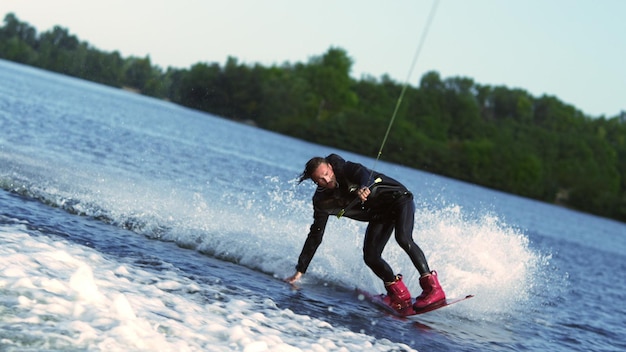 Mann rast mit Wakeboard am Waldsee entlang und berührt mit der Hand die Wasseroberfläche