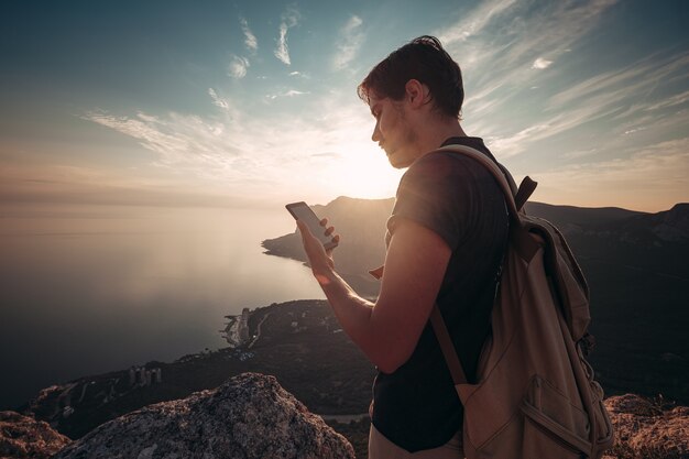 Mann Rucksacktourist mit Smartphone entspannen auf Berggipfel allein reisen. Lifestyle Aktivurlaub Moderne Technologie Millennials Konzept. Herrlicher Tagesanbruch. Mock-up-Text.