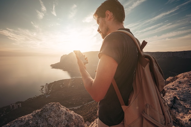 Mann Rucksacktourist mit Smartphone entspannen auf Berggipfel allein reisen. Lifestyle Aktivurlaub Moderne Technologie Millennials Konzept. Herrlicher Tagesanbruch. Mock-up-Text.