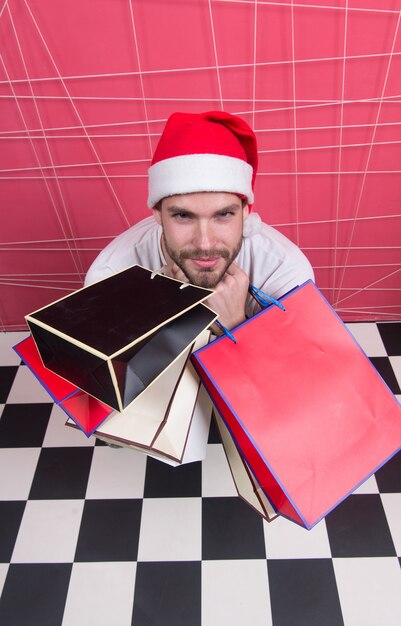 Mann Sankt halten Taschen auf schwarz-weiß kariertem Boden. Shopper im roten Hut mit Papiertüten, Ansicht von oben. Weihnachten, Neujahrsgeschenke. Winterschlussverkauf, Einkaufskonzept. Vorbereitung und Feier der Feiertage.