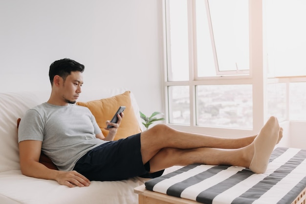 Mann schaut auf sein Handy, während er sich auf dem Sofa in seinem Wohnzimmer entspannt