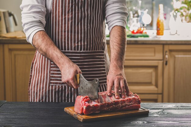 Mann schneidet frisches Fleisch auf einem Holzbrett in der heimischen Küche. Ein Mann in einer gestreiften Schürze mit einem großen Messer in der Hand