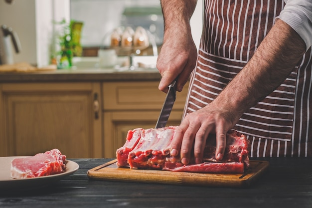 Mann schneidet frisches Fleisch auf einem Holzbrett in der heimischen Küche. Ein Mann in einer gestreiften Schürze mit einem großen Messer in der Hand