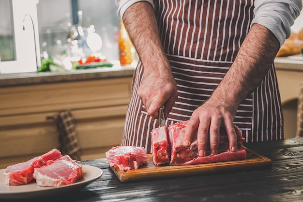 Mann schneidet frisches Rindfleisch auf einem Holzbrett in der heimischen Küche. Ein Mann in einer gestreiften Schürze mit einem großen Messer in der Hand