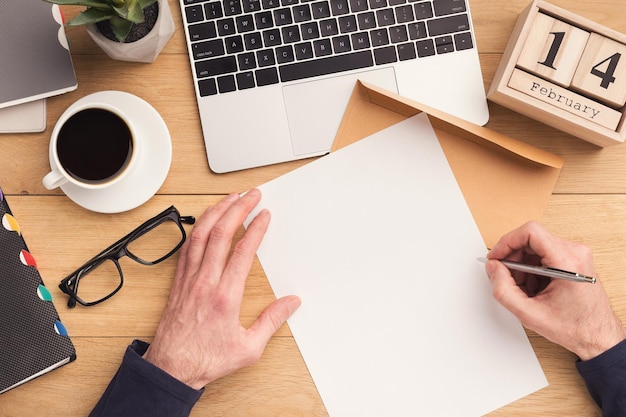 Mann schreibt Brief, während er am Laptop arbeitet und Kaffee auf Vintage-Holztisch trinkt, Ansicht von oben. Männlicher Arbeitsplatz mit Kakteen, ewigem Kalender und Brille