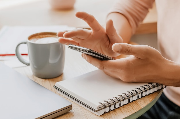 Mann schreibt Geschäftsinformationen in Notizbuch am Arbeitsplatz im Büro mit Smartphone