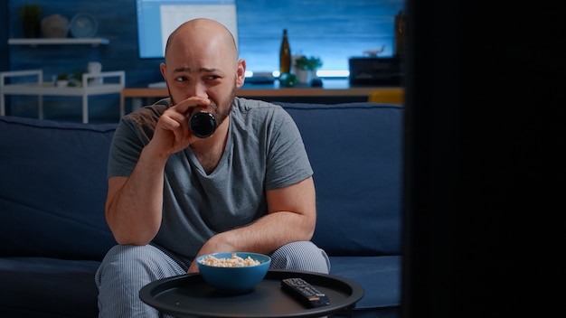 Mann sitzt allein auf der gemütlichen Couch im Wohnzimmer und isst Popcorn, trinkt Bier?