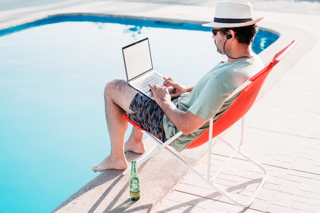 Mann sitzt allein mit einem Laptop auf einem Liegestuhl am Pool