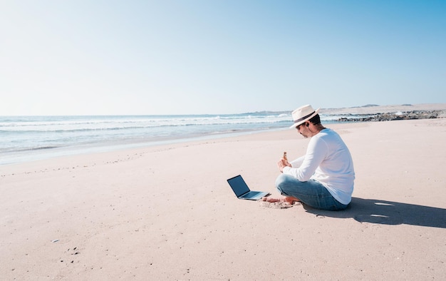 Mann sitzt am Strand mit einem Laptop allein, macht Telearbeit oder Fernarbeit und trinkt Bier