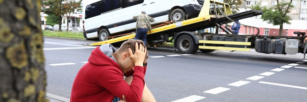Mann sitzt am Straßenrand und hält seinen Kopf mit den Händen neben dem zerstörten Auto nach einem Autounfall. Verkehrsverletzungskonzept