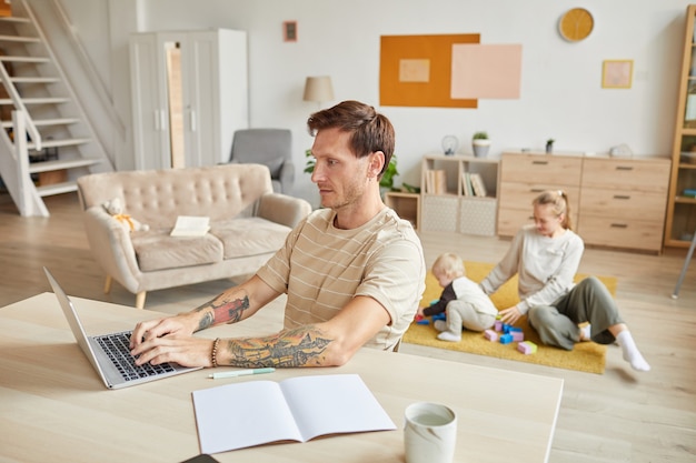 Mann sitzt am Tisch und arbeitet am Laptop mit seiner Familie, die zu Hause spielt