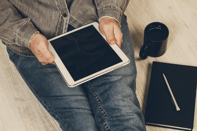 Mann sitzt auf dem Boden mit einer Tasse Kaffee und arbeitet mit einem Tablet