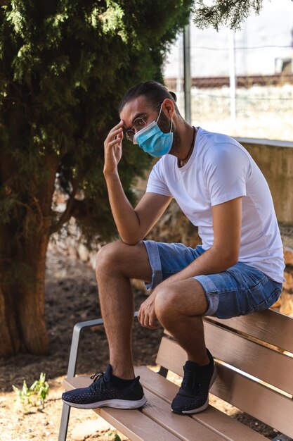 Mann sitzt auf einer Bank mit Maske, weißem Hemd und Jeans