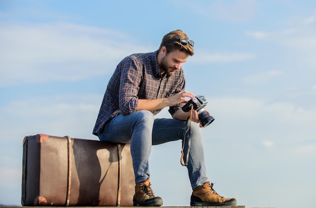 Mann sitzt auf Koffer Reiseblogger Hübscher Kerl Reisende Retro-Kamera Reisen mit Gepäck Schießen vlog Urlaubszeit Kerl im Freien mit Vintage-Koffer Fotojournalist Konzept Reiseblog