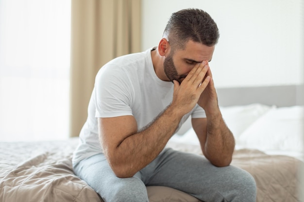 Mann sitzt mit den Händen auf dem Gesicht im Zimmer auf dem Bett
