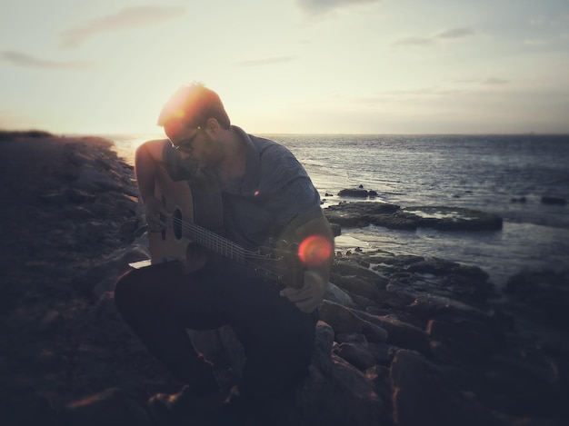 Mann spielt Gitarre, während er bei Sonnenuntergang gegen das Meer sitzt