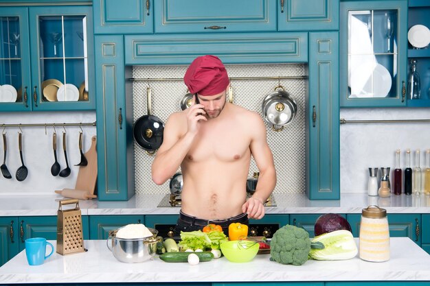 Mann spricht am Handy in der Küche Koch in Kochmütze mit sexy Oberkörper am Tisch Vegetarisches Menü und gesunde Ernährung Zubereitung von Speisen und Kochrezepte Kommunikation und neue Technologie