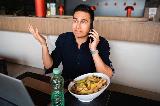 Mann spricht am Telefon und sitzt am Tisch, während er mit dem Computer gesund zu Mittag isst Männliche erhobene Arme