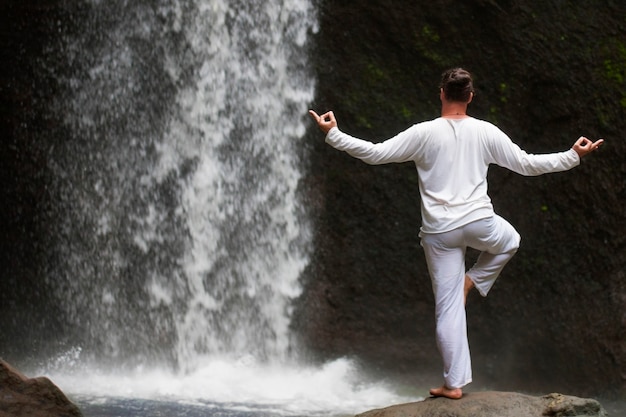 Mann steht im Meditationsyoga auf Felsen am Wasserfall im tropischen Bali